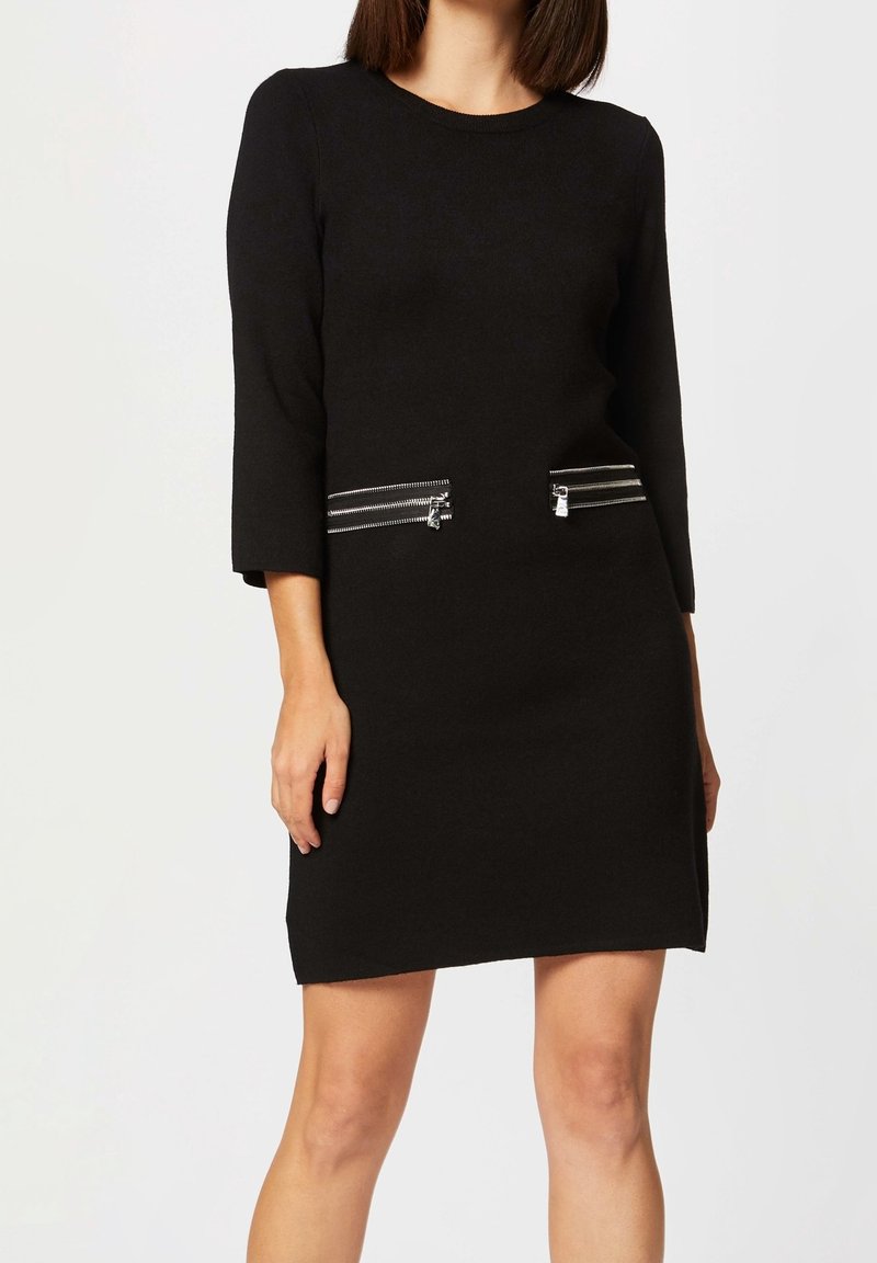 Robe noire à manches trois-quarts, avec deux poches à glissière argentées sur le devant. Texture lisse et design à coupe droite.