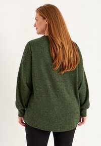 Groene gestructureerde trui met een ronde zoom en lange, iets opgeblazen mouwen. Model staat met de rug naar de camera, gecombineerd met zwarte leggings.