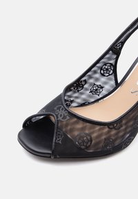 Μαύρο ανοικτό παπούτσι τύπου slingback με διάφανα τοιχώματα από δίχτυ με λουλουδένια μοτίβα και χαμηλό τακούνι σε λευκό φόντο.