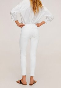 Jeans blancos ajustados con cintura alta, que cuentan con bolsillos traseros y aberturas en los tobillos, combinados con una camisa blanca suelta y sandalias.