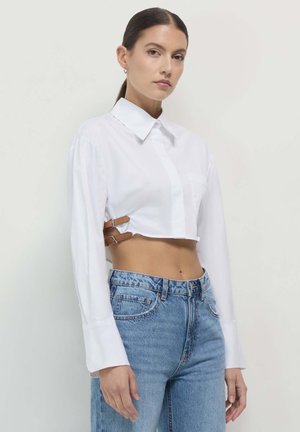 White cropped blouse with classic collar, long sleeves, and two brown leather straps at the waist. Paired with light blue high-waisted jeans.