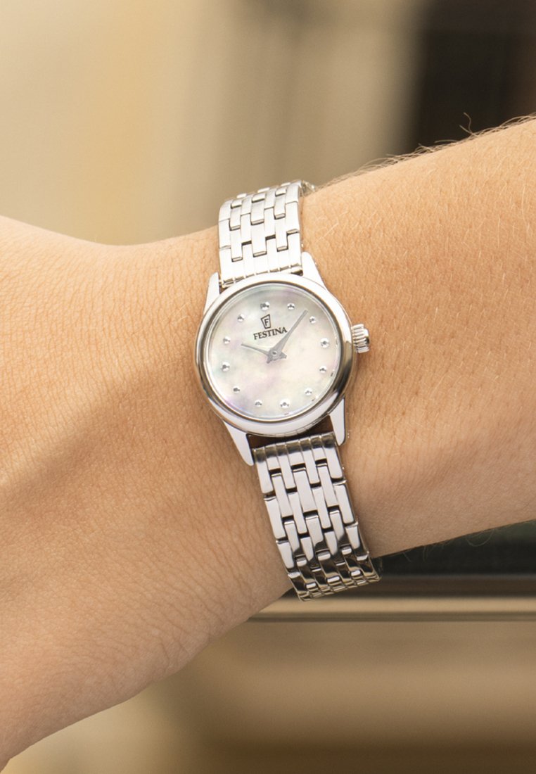 Reloj de pulsera Festina plateado con esfera blanca y pequeños marcadores de hora de cristal en la muñeca de una persona.