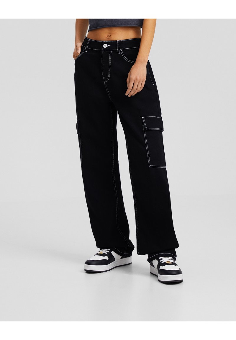 Pantalones cargo negros con costuras blancas, corte holgado, bolsillos laterales y cierre de botón. Combinados con zapatillas negras y blancas.