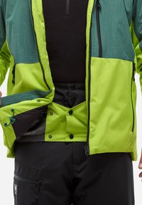 Veste imperméable verte et noire avec un motif en grille, poches zippées et cordon de serrage ajustable. Associée à un pantalon noir avec des boutons et des zippers.