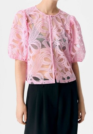 Pink lace short-sleeve blouse with floral patterns and buttons down the front, worn over a black top and paired with black pants.