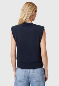Marineblauw mouwloos gebreid topje met een ronde hals, geribbelde zoom en bijzondere schouderdetails, gecombineerd met lichtblauwe hooggetailleerde jeans.