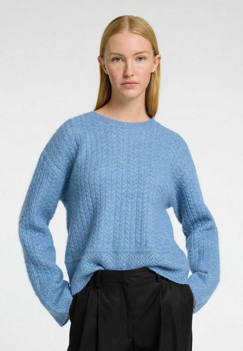 Lyseblå strikket sweater med kabelmønster, rund halsudskæring og let oversized pasform, parret med sorte bukser. Blød tekstur.