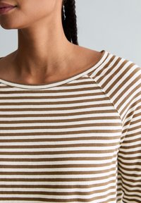 Frau trägt ein braun-weiß gestreiftes Langarmshirt mit rundem Ausschnitt, dargestellt von den Schultern bis zum Hals.