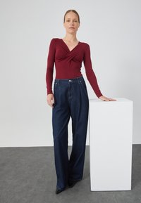 Top avvolgente bordeaux a maniche lunghe abbinato a jeans scuri in denim a vita alta e morbidi. Il modello si trova accanto a un piedistallo bianco.