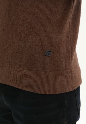 Brūns adīts džemperis ar teksturētu audumu, ar ribotu apakšmalu, kurā ir neliels melns logo uzlīme sānos. Nēsāts ar tumšām biksēm.