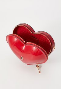 Red heart-shaped bag with a glossy finish, gold zipper, and key charm detail. Interior features smooth lining with logo print.