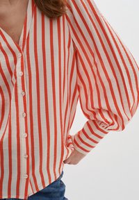 Chemise à manches longues avec des rayures verticales rouges et blanches, en tissu doux et texturé, et manches bouffantes, partiellement rentrées dans un jean bleu.