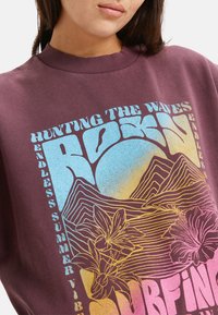 Bordeaux sweatshirt met geribbelde halslijn, voorzien van een kleurrijk grafisch ontwerp met tekst en bloemelementen in blauwe, gele en roze tinten.