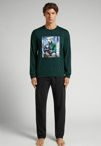 Groene sweatshirt met lange mouwen met een kleurrafische afbeelding van een hond in een jas. Gepaarde met zwarte broek, tegen een neutrale achtergrond.