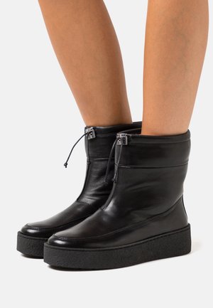 Black mid-calf leather boots with thick textured platform soles and adjustable drawstring closures worn on bare legs.