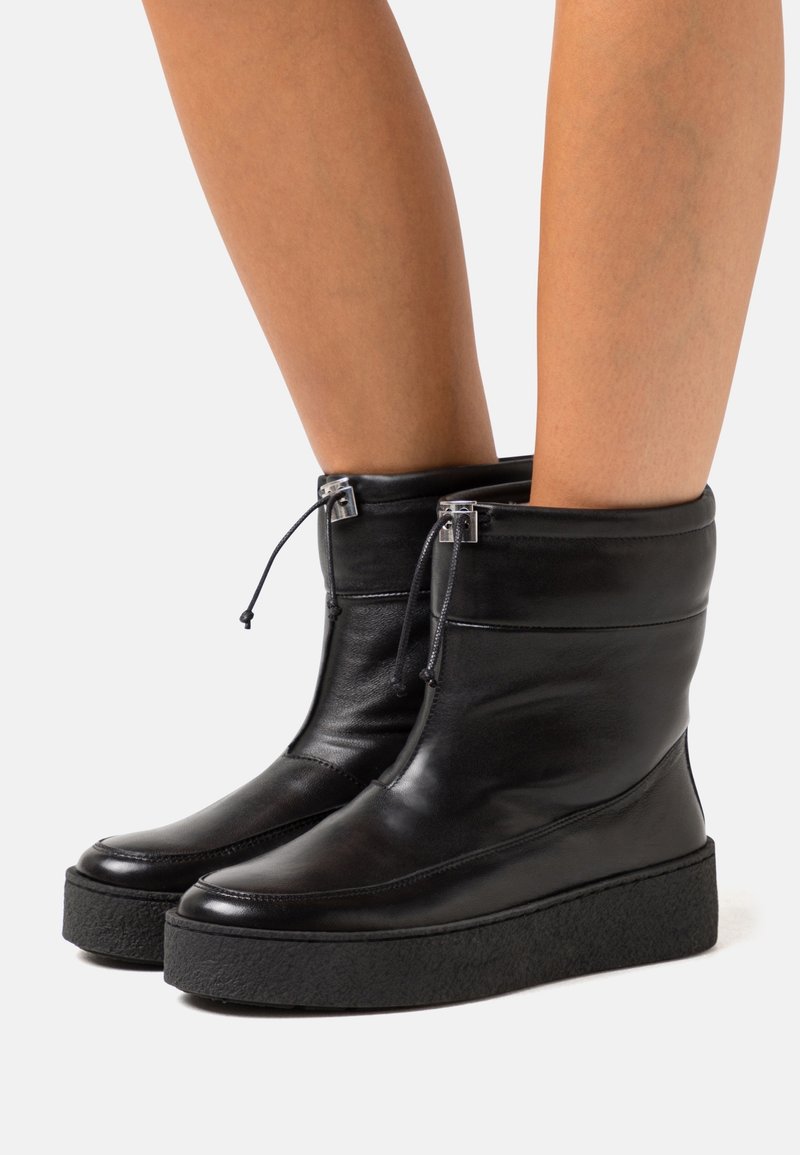 Bottes noires en cuir jusqu'au milieu du mollet avec semelles plates-formes épaisses et texturées et fermetures à cordon ajustables portées sur des jambes nues.