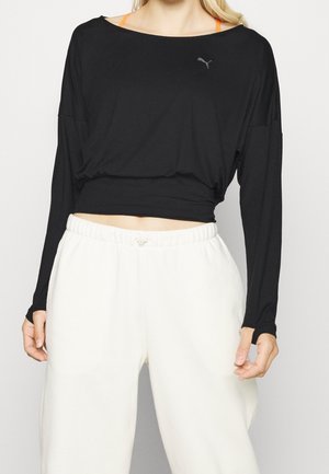 Vrouw die een zwart cropped top met lange mouwen draagt over een oranje sportbeha en losse witte joggingbroek, staand tegen een effen achtergrond.