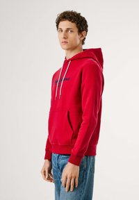 Rode hoodie van katoen met een kangoeroezak, koordjes en een logo op de borst. Gekleed met blauwe jeans met een casual pasvorm.