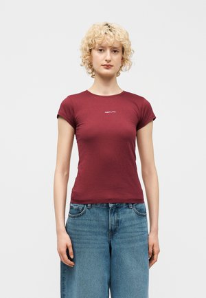 Osoba o blond kręconych włosach, ubrana w dopasowany bordowy t-shirt z małym białym logo oraz luźne niebieskie dżinsy, stojąca na tle jednolitego tła.