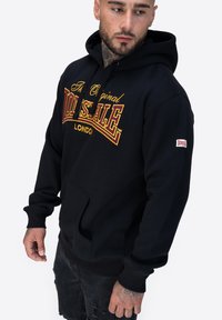 Hoodie nero in cotone, con tasca frontale, polsini a coste e stampa grafica in oro e bordeaux. Etichetta del marchio sulla manica.