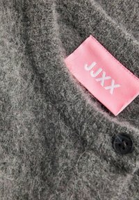 Šedá vlněná tkanina s chlupatou texturou, na které je růžová nálepka s nápisem "JUXX" bílou barvou, a černý detail knoflíku na límci.