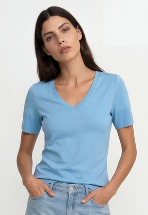 Frau mit langen dunklen Haaren, die ein hellblaues Kurzarm-V-Ausschnittshirt und hellblaue Jeans trägt, steht vor einem schlichten weißen Hintergrund.