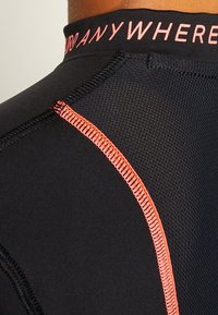 Schwarzes Sportshirt mit enganliegendem Design, aus strukturiertem Material, kontrastierenden pinken Nähten und einem "ANYWHERE"-Label am Kragen.