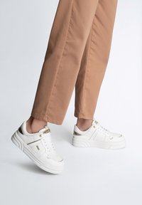 Sneaker bianchi in pelle testurizzata, con dettagli dorati e una suola in gomma spessa, abbinati a pantaloni beige dritti.