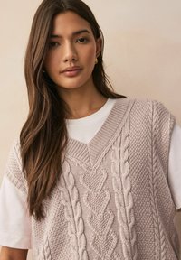 Vest tricotat în roz deschis, cu modele de cabluri texturate, purtat peste un tricou alb. Are un design cu guler în V și un material moale și comod.