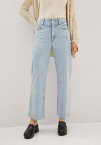 Jean bleu clair taille haute à jambes droites avec ourlet effiloché et cinq poches, associé à des sabots noirs ornés de clous dorés.