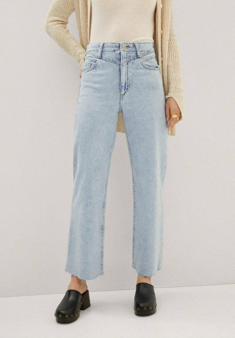 Jean bleu clair taille haute à jambes droites avec ourlet effiloché et cinq poches, associé à des sabots noirs ornés de clous dorés.