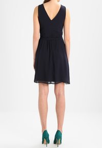 Marinblå ärmlös klänning med V-rygg, insvängd midja och plisserad kjol. Matchas med turkosa högklackade skor. Slät tygstruktur.