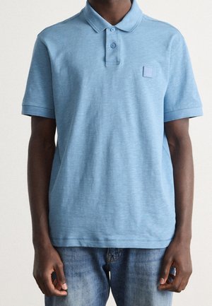 Mand iført en lyseblå kortærmet polo med to-knaps åbning og denimjeans, stående mod en ensfarvet baggrund.