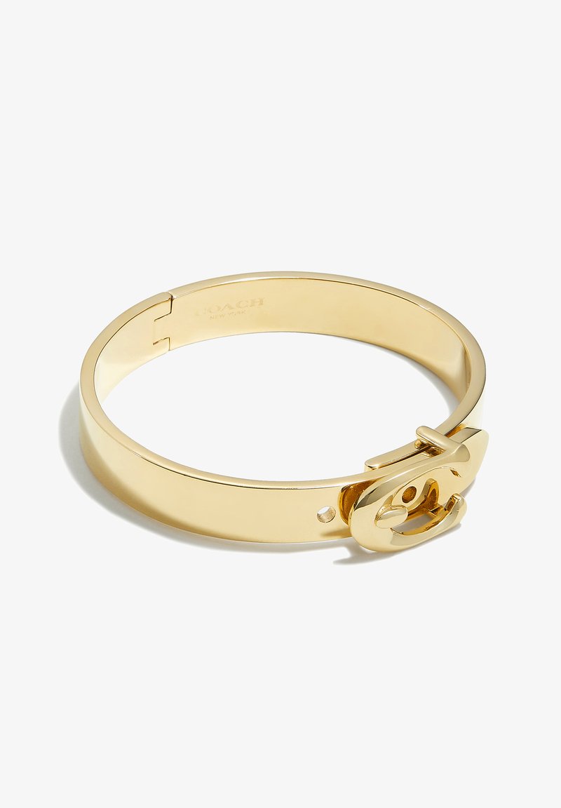 Pulsera de bangle de metal en tono dorado con un acabado suave, que presenta un cierre prominente diseñado en forma de un logotipo estilizado.