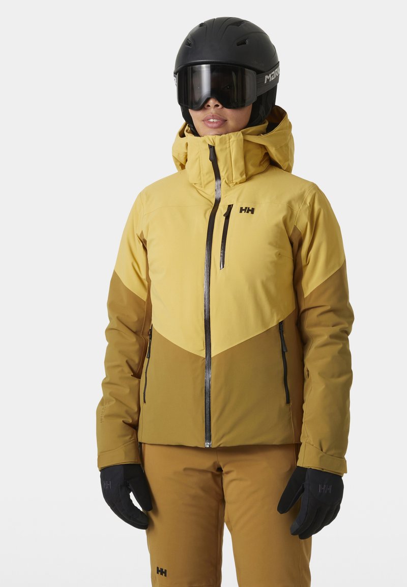 Geel en bruine skijas met een capuchon, voorzien van zwarte ritsen en een logo. Combinatie met bijpassende broek en zwarte handschoenen voor sneeuwsporten.