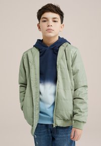 Svetlo zeleni pufasti plašč z zadrgo spredaj, v kombinaciji z mornarsko modrim kapuco s sweatshirtom, ki ima ombre efekt. Obučen s strganimi jeansi.