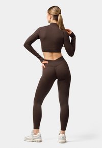 Brauner, langärmeliger Crop-Top und hochgeschnittene Leggings, beide mit gerippter Textur. Das Outfit zeichnet sich durch ein tailliertes Design und minimale Nahtdetails aus.
