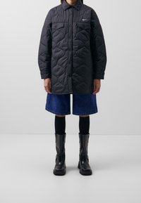 Svart vadderad jacka med knappar framtill, två bröstfickor och logotypdetalj, tillsammans med blå denimshorts och svarta knähöga stövlar.