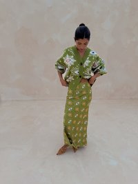 Cardigan vert en maille avec motifs floraux et d'œil, associé à une longue jupe verte avec un motif similaire. Chaussures marron pointues ornées de clous.