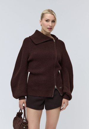Donna con capelli biondi che indossa un maglione marrone scuro con cerniera e maniche a sbuffo e pantaloncini abbinati, che tiene in mano una piccola borsa marrone.