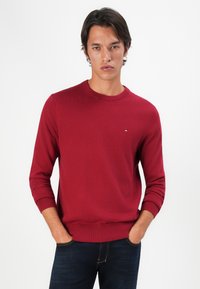 Pull rouge col rond à manches longues, texture tricotée, avec des poignets et un ourlet côtelés, arborant un petit logo sur la poitrine. Porté avec un jean foncé.