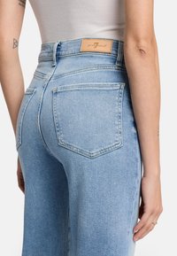 Jean en denim bleu clair à taille haute, avec deux poches arrières et une étiquette en cuir sur la ceinture. Texture lisse.