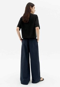 Schwarzes, cropped T-Shirt mit kurzen Ärmeln, kombiniert mit weiten, marineblauen Hosen, von hinten auf einem hellen Hintergrund präsentiert.