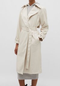 Trench-coat beige avec une taille ceinturée, un grand col et des manches retroussées. Tissu lisse, longueur jusqu'aux genoux, avec poches des deux côtés ; superposé sur un sweat à capuche gris.