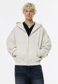 Ein off-white Zip-Hoodie mit lockerer Passform, großen Fronttaschen und fransigen Säumen, kombiniert mit einer dunklen Jeans.