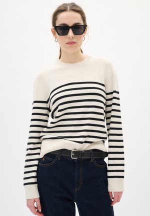 Maglione beige con righe orizzontali nere, scollatura rotonda e polsini a coste, abbinato a jeans blu scuro e una cintura nera. Occhiali da sole neri.