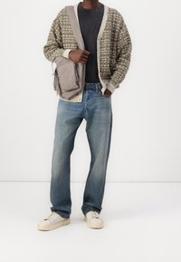 Stickad kofta i olivgrönt och krämfärg, svart t-shirt, ljusblå jeans, krämfärgade sneakers och grå axelremsväska med dragkedjor och fickor.