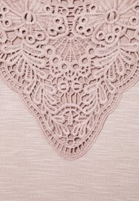 Rosa tyg med ett intrikat spetsmönster i mitten. Spetsen har blommiga och cirkulära motiv, vilket ger textur till den släta bakgrunden.