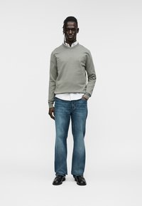 Πουλόβερ σε ανοιχτό γκρι βαμβακερό μείγμα, στρογγυλή λαιμόκοψη· φορεμένο πάνω από ένα λευκό πουκάμισο με κουμπιά. Τζιν παντελόνι σε μπλε αποχρώσεις και μαύρα δερμάτινα παπούτσια ολοκληρώνουν το σύνολο.