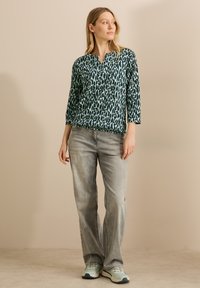 Långärmad topp i teal med ett svart abstrakt mönster, tillsammans med ljusgrå jeans och blå sneakers. Toppen har en V-ringning.